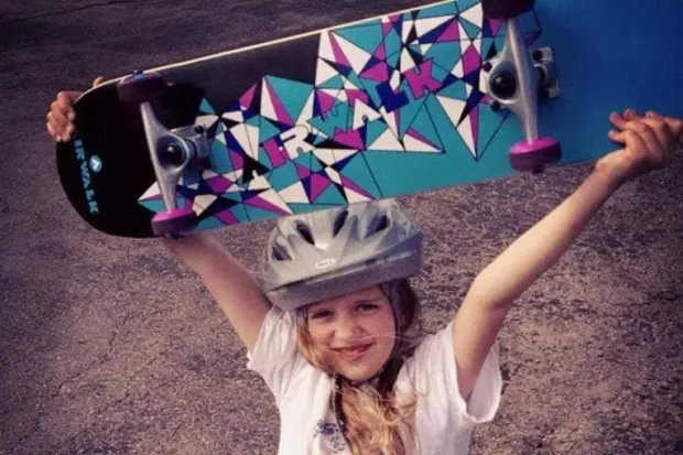 Young girl wearing helmet holding skateboard over her head 