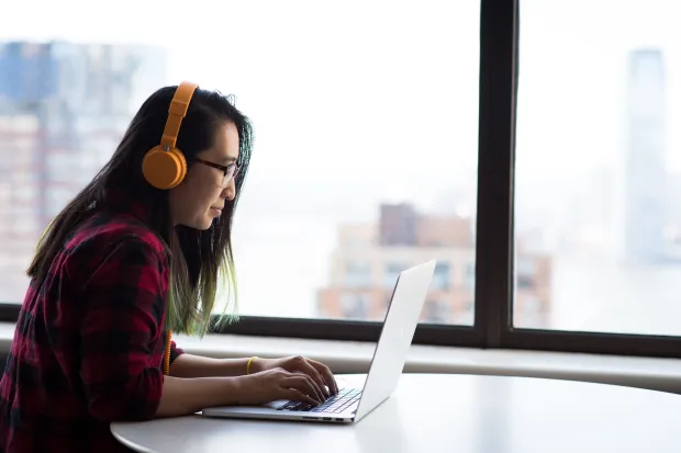 A girl wearing headphones sitting at a laptop near a window