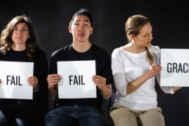 Three young people sitting holding signs. Two wearing black T-shirts hold up signs with "Fail" written on them. The third woman is wearing white holding a sign that says "Grace."