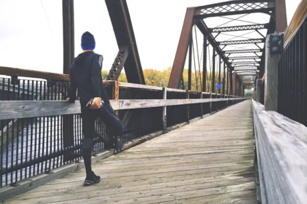 Man in jogging clothes stretching leg, leaning on railing on bridge