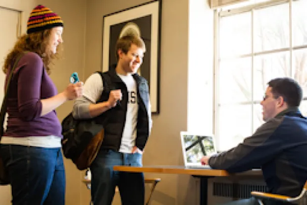 Male and female student walking up to campus minister at table with computer