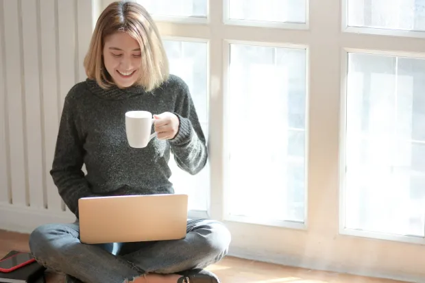 a girl seated cross-legged by a sunny window, holding a mug and using her laptop