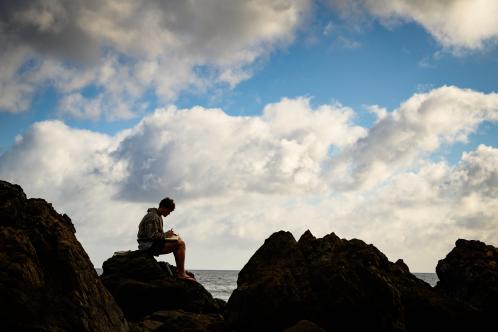 student sitting on a rock near the water with the backdrop of a blue sky with white clouds