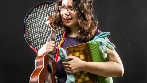 Female college student holding guitar, tennis racket, chessboard, books, and blanket looking overwhelmed