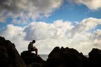 student sitting on a rock near the water with the backdrop of a blue sky with white clouds
