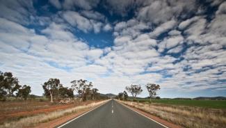 Highway running through rugged landscape with scattered trees and swathes of white clouds