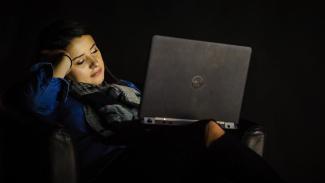 Woman in dark room with laptop glowing on face, looking bored