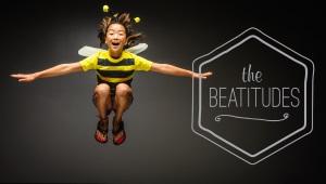 Young child in bee costume in midair, grinning, with the words "The Beatitudes" in the background.