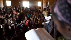 An African woman reading a Bible to a crowded room of listeners