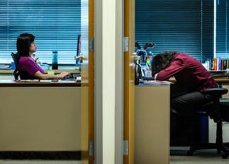 Two offices side by side with doors open. One woman is typing at her computer while the woman in the next office has her head down on the desk.