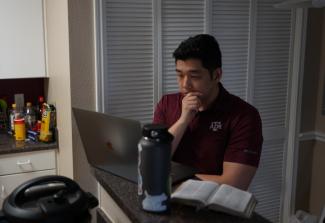 Student reflecting on Scripture, standing at kitchen counter with laptop