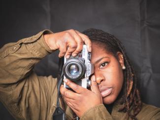 A young black woman holding a camera over her right eye