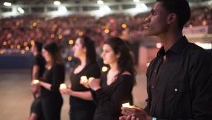 Group of students wearing black holding candles with much larger group in the background in stadium seating