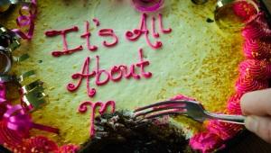 Closeup of pink and yellow cake with "It's All About Me" written in icing. Part of the "Me" has been eaten with a hand and fork poised for more.