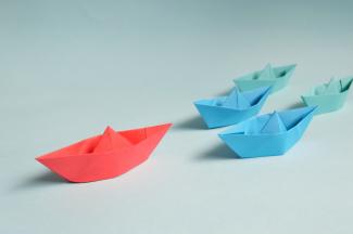 Group of origami boats. A red one is in the lead with blue and green ones trailing after.