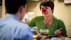 Middle-aged woman wearing red clown nose talking to man across table