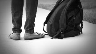Black and white closeup of person's legs standing on sidewalk next to backpack