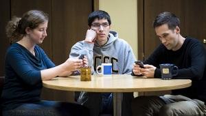 Three people sitting around table. Two staring at phones. One looking bored.