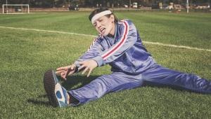 Person on soccer field in bright purple tracksuit grimacing as she stretches