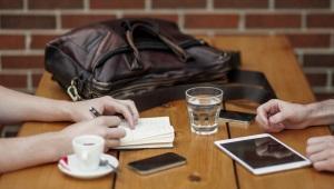 two pairs of hands resting on a wooden table with cups and an iPad on it.