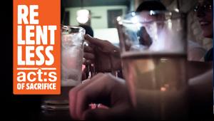 Closeup of hand next to glass with the label: Relentless Acts of Sacrifice