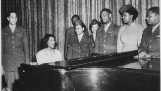 a photo from the 1940's of Black people standing around a piano and singing