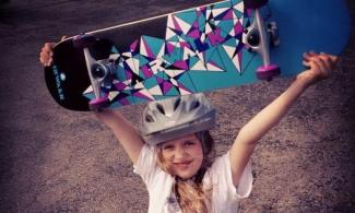 Young girl wearing helmet holding skateboard over her head 