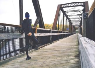 Man in jogging clothes stretching leg, leaning on railing on bridge