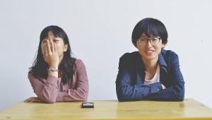 Young couple sitting beside one another. The man is smiling, and the woman is face-palming. 