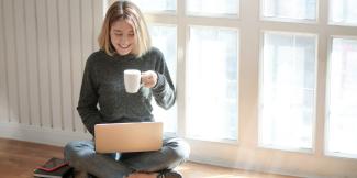 a girl seated cross-legged by a sunny window, holding a mug and using her laptop