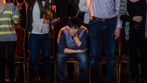 a sad-looking man sitting down in a crowd of standing people