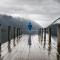 A person standing a long at the end of a rainy dock, facing a lake and distant mountains