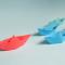 Group of origami boats. A red one is in the lead with blue and green ones trailing after.