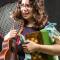 Female college student holding guitar, tennis racket, chessboard, books, and blanket looking overwhelmed