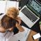 Woman looking stressed, holding her head surrounded by computer and notebooks