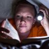 Young woman reading book under covers with flashlight