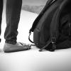 Black and white closeup of person's legs standing on sidewalk next to backpack