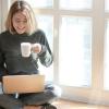 a girl seated cross-legged by a sunny window, holding a mug and using her laptop